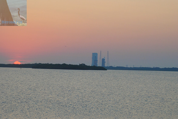 An outdoor, sunrise photograph of the Sun, made to look pink in the early morning, which has just risen halfway above the horizon. The sky is pink and orange at the top of the image, with more gray clouds near the middle, where trees and brush cover the horizon. Near the middle of the image is a tall white tower, to the right of which are two tall, truss-work towers and a shorter, gray, truss-work structure with connections leading to an orange-colored rocket.