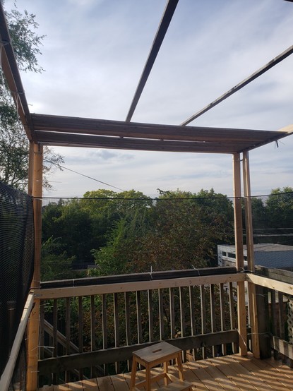 A rudimentary sukkah under construction: wooden frame of 2×4 boards 8 feet square, lower walls being the existing wooden porch fence, upper walls black plastic garden trellis mesh, and a minimal amount of sekhakh (sukkah roofing) in place, of 1×2 wooden boards.