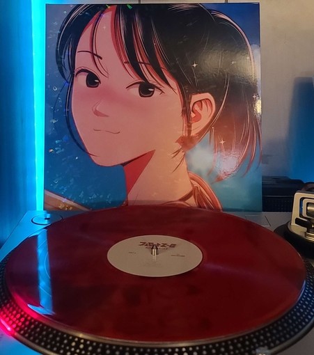 A transparent red vinyl record sits on a turntable. Behind the turntable, a vinyl album outer sleeve is displayed. The front cover shows an anime girl smiling and looking at the camera with a blue sky and sun behind her