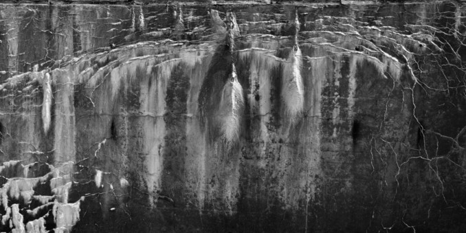 This wide format digital photograph shows a somewhat mustache shaped crack in a concrete wall, weeping bright precipitates over a darker background of the wall itself.