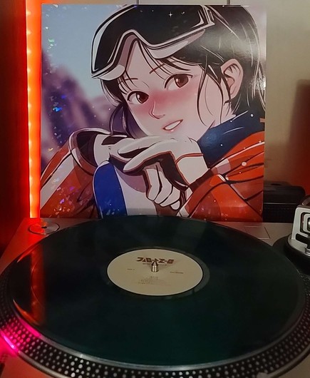 A green translucent vinyl record sits on a turntable. Behind the turntable, a vinyl album outer sleeve is displayed. The front cover shows an anime girl in a ski outfit and goggles looking at the camera and smiling
