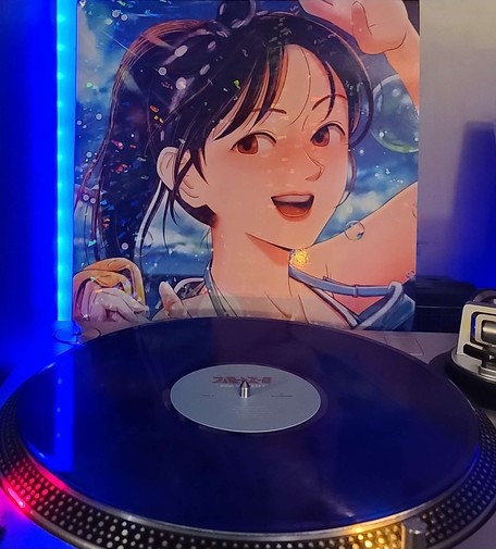 A Blue clear vinyl record sits on a turntable. Behind the turntable, a vinyl album outer sleeve is displayed. The front cover shows an anime girl w/ a ponytail smiling with the ocean and blue sky behind her.