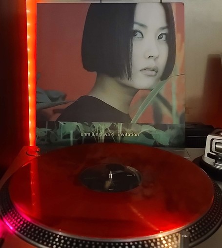 A red transparent vinyl record sits on a turntable. Behind the turntable, a vinyl album outer sleeve is displayed. The front cover shows Uhm Jung Hwa looking to her right with tall plants around her