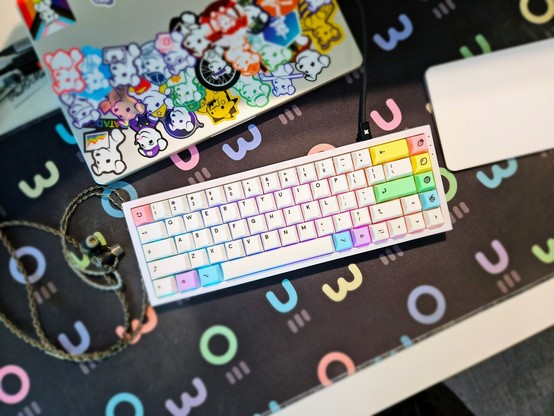 Top down photo of a small form factor, white mechanical keyboard with an Apple Trackpad beside it and a Macbook Pro on the edge of the shot with many stickers on it featuring the Datadog logo in different forms.