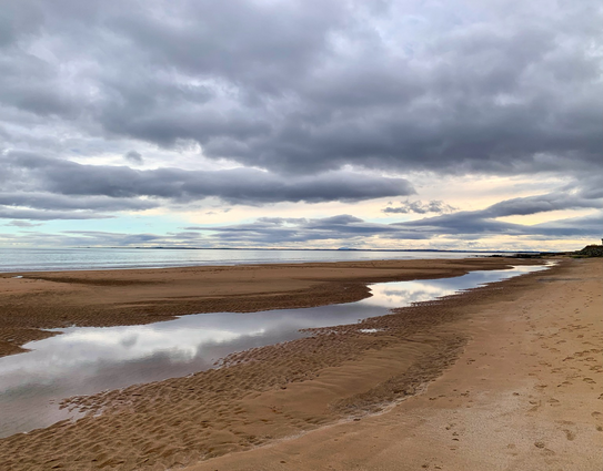 A beach photo. Banks of grey cloud with touches of white and a slice of pale blue,left. Sea a sliver of silver. Dark sand newly wet, a timeline, puddled with water reflects the clouds.