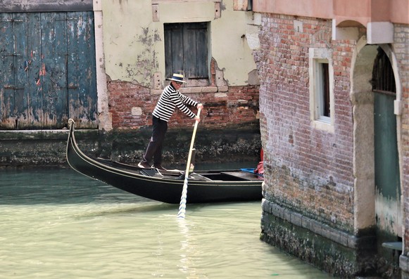 A narrow backwater in Venice, with crumbling, brick walls, and wooden dooors and windows. A smiling gondolier is rowing his boat past a side turning.