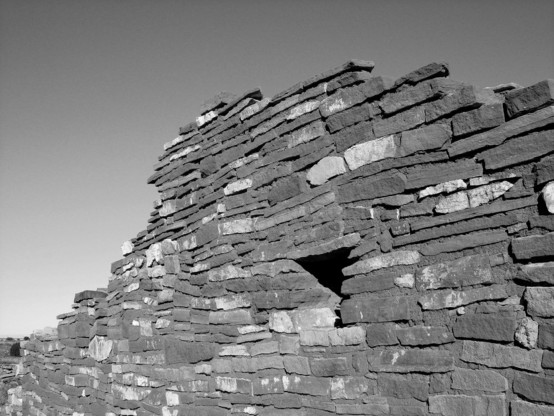 Black and white photo of the remains of a wall composed of flat stones and mortar, which features a small, square window partway up
