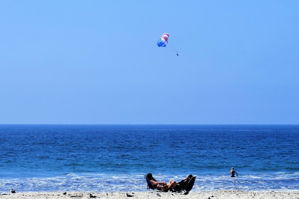Two people in beach chairs close to the water.  A man entering the surf and a paraglider over the water.