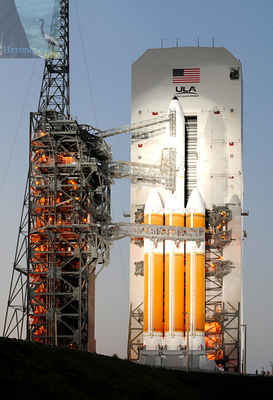 An outdoor, evening photograph of a rocket sitting on its launch pad with umbilical cables connected. The rocket has 3 orange cylindrical boosters side-by-side with the center booster carrying the white upper stage and fairing. Behind the rocket is a white tower with an opening that can hold the rocket. On the right side of the image is a truss tower with three truss connections to the rocket. Behind it is a tall, narrow truss tower extending beyond the top of the image.