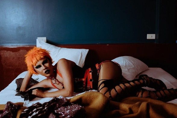 Personal with orange hair and a collar laying seductively on bed wearing read leather straps and fenestrated black boots.