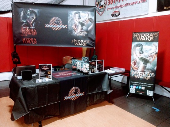 Image shows the Creature Author booth setup for Firghtreads Book Festival. Showcasing the Hydra's Wake, and Hydra Tower cover art, signage about prices, a signing mat, Paperbacks and Hardcovers, the Creature Author Logo on the tablecloth.