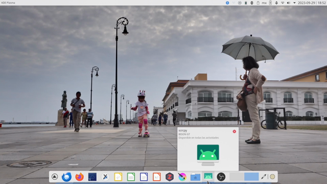 Escritorio KDE Plasma, con paneles claros, uno superior para el control de ventanas, menú global, bandeja del sistema y reloj; otro inferior (flotante) como dock.
La imagen de fondo es una foto del Malecón de Veracruz, donde se ve a una niña viniendo hacia la cámara en una patineta, a una lado, una señora con sombrilla la observa.
Desde el dock, se pulsa sobre un icono que hace referencia a Android, mostrando la ventana del programa scrcpy, que permite ver e interactuar con la pantalla de celulares Android. El fondo de pantalla del móvil, es una foto de la misma escena, pero la niña está más cerca de la cámara y en una pose más dinámica, guardando el equilibrio, sobre su patineta.
La ventana de scrcpy es minimizada y mostrada varias veces, en forma semejante a una animación tradicional por superposición de imágenes.