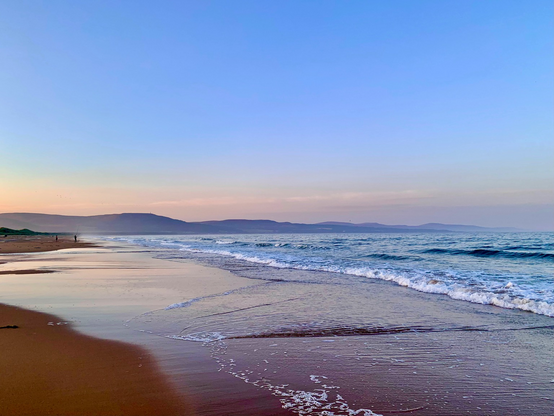 A photo of a beach at sunset. Clear pale blue sky too with a pinky peachy glow mid photo. Misty blue grey hills underneath. Sea to the right beach to the left. Wa yes breaking. People silhouetted in the distance, left, centre. Clouds ,reflected on the shore.