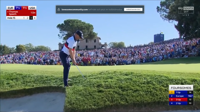 ein amerikanischer Golfspieler steht an einer Bunkerkante im dichten Gras und versucht seinen Ball auf das Green zu spielen, um den dann dort ins Loch zu putten.
Im Hintergrund an der Greenkante stehen Hunderte Zuschauer und Fans