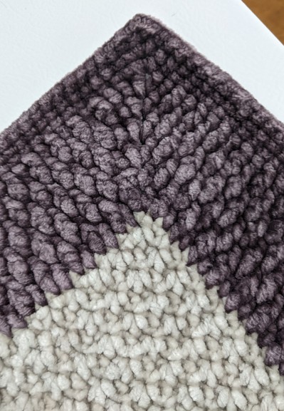 The corner of a purple and white crocheted baby blanket. A close-up intended to show off the various patterns created by the different stitches.
