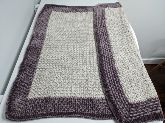 A white (almost silver) crocheted baby blanket with a deep purple border. One side is folded over to show the slight variation in the stitch pattern on the back.
