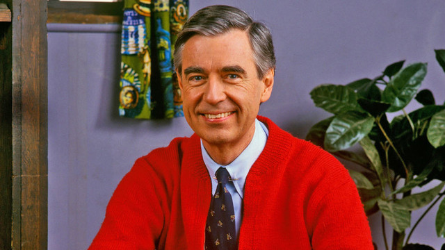 A picture of Fred Rogers looking into the camera and smiling on the set of Mr. Rogers' Neighborhood. In this picture he's an older white man with dark grey hair. He's wearing a red sweater, a light blue button up shirt, and a black tie.