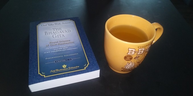 An orange mug filled with tea next to a copy of the Bhagavad Gita with Paramahansa Yogananda's commentary.