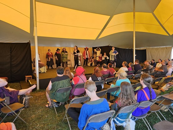 Numerous players on a raised wooden stage before an audience in a pavilion tent during a medieval camping perform a long-form improv comedy show.