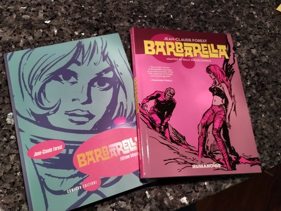Two graphic novels on a granite surface. One cober has a with a teal background and an image of Barbarella's face, the other with a rose background and Barbarella backed up against a rock with a male companion climbing towards her.