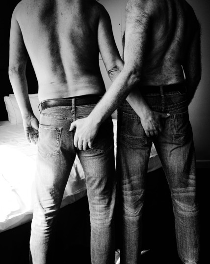 Two men stand with their backs to the camera.  They are topless and wearing jeans.  They each have a hand on the others arse .