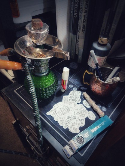 A hookah pipe with a lit coal on a mat covered in cut-out speech bubbles and a gluestick, including one that says "SHUT YOUR MOUTH, GUTTER SNIPE!" in large dramatic letters. A bundle of sage, a large pour bottle of black ink, and two lead refills are visible in the foreground.