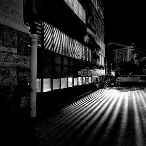 Lateral view of a house facade in the dark of the evening. The picture shows a continuous window front on three levels next to a vertical rain gutter. The light of the illuminated rooms falls through the horizontal slats of blinds and casts a striped pattern on the path in front of the building, on which a bicycle with the sign "STADT THEATER" is parked.