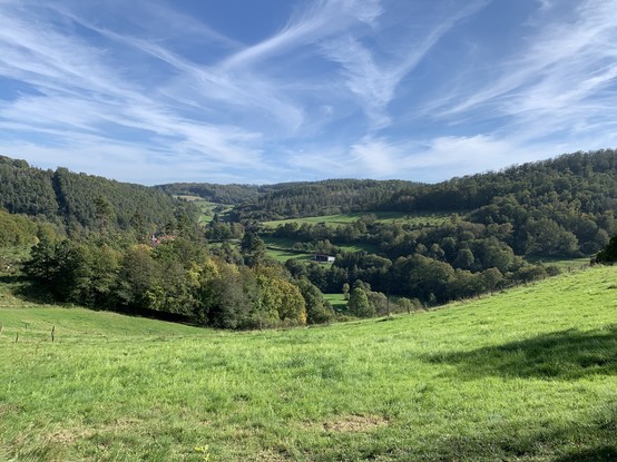 View of green lawns and forests down from a hill, under a blue sky with wispy clouds. Photo was taken on a hike in national park Kellerwald-Edersee in the state of Hesse, Germany.