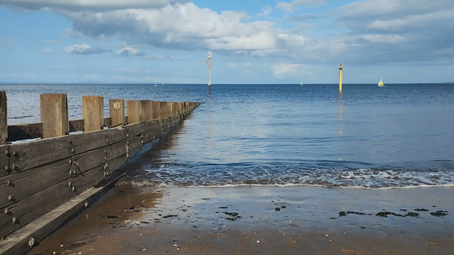 Looking down the sloping length of the wooden groyne on Portobello Beach, as it descends into the Firth of Forth, as gentle waves lap the shore, then turning right to take in more of the beach