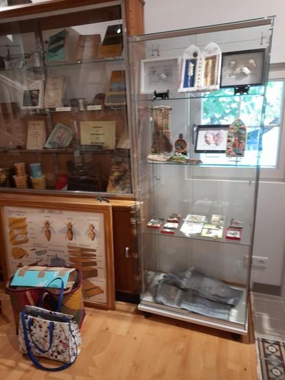 A glass case next to an old display cabinet in the museum. In the cabinet, historical accessories for beekeeping. In the showcase, my Biodiversity Shrines and at the bottom a jeans for visible mending. A huge handbag and an even bigger basket bag with packaging waste are standing around - only staff members are allowed to place such things.