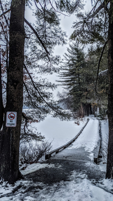 Walking along a dirt and gravel footpath beside a lake. Straight ahead, we are approaching a rather long wooden footbridge across the neck of a small cove. Groves of tall pines and hemlocks grace the shores on both sides of the bridge. The lake surface is frozen. A dusting of snow covers the ice and the ground and has also been caught in some of the tree branches. A very light snow is falling from the overcast sky, slightly ghosting the far shore of the lake along the horizon.