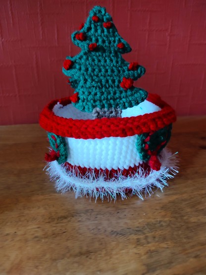 A photo of a Christmas crochet treat basket showing one of the two green Christmas trees that make up the handle from the front. Red bobbles are visible on the tree. The red rim of the basket is clearly visible and two of the little green wreaths can be seen on either side on the white middle part of the basket in profile. There is a little later of white tinsel yarn that adorns the bottom of the basket.