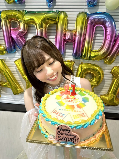 Sakuri posing in her stage outfit, smiling looking at her birthday cake with baloons in the background saying Happy Birthday Sayuri.