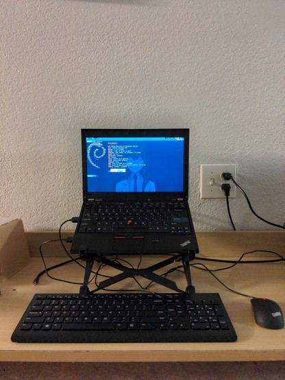 A thinkpad X230 on a nextstand with a lenovo keyboard and a thinkpad mouse on a motel 6 desk