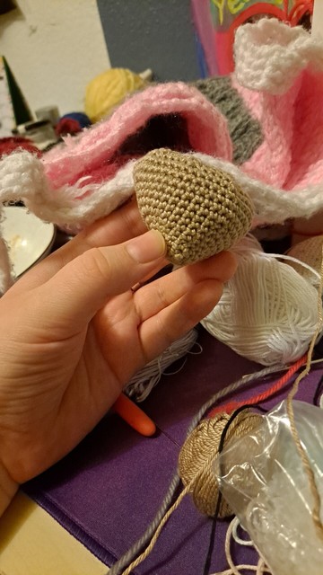 A beige shape crocheted with fine yarn and stuffed. It's a bit like a funky strawberry shape. A hand holds it over a cluttered table.