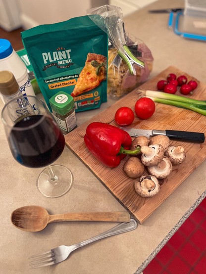 Wooden cutting board with mushrooms, tomatoes, spring onions, radishes, red bell pepper, next to salt, olive oil, pasta, grated vegan cheddar cheese and a glass of red wine. Red tiles visible on the right bottom corner.