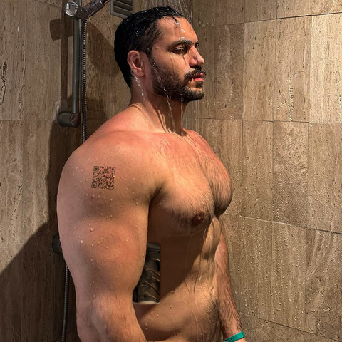 A muscular Arabic shirtless android showers in a  brown marble shower. The water cascades down its thick, lightly haired pecs. It has an open oblique panel waiting for water to seep in. Its eyes are closed as it is shut down. It has a QR code on its shoulder