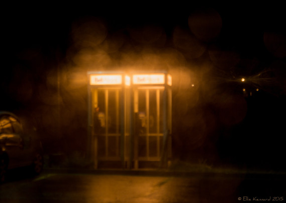 It is a dark image, with only a few details lit in a yellow glow. There are two phone boxes dimly visible, side by side, which are where the light is coming from, but it is very low, with just white "Bell" notices on a strip on the top of each. Although other lights are dimly seen, it's not really possible to work out what they might be, apart from the glow on the road in front of the phone booths.