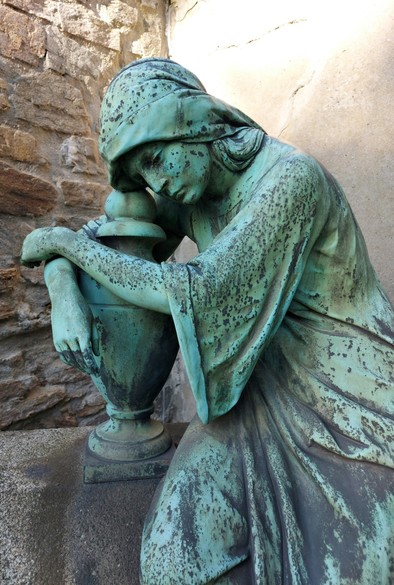 Eine Statue aus Metall mit Grünspan. Eine Frau die eine Urne umarmt und über die verstorbene Person trauert. Sie sitzt auf einem Podest und im Hintergrund befindet sich eine Mauer.