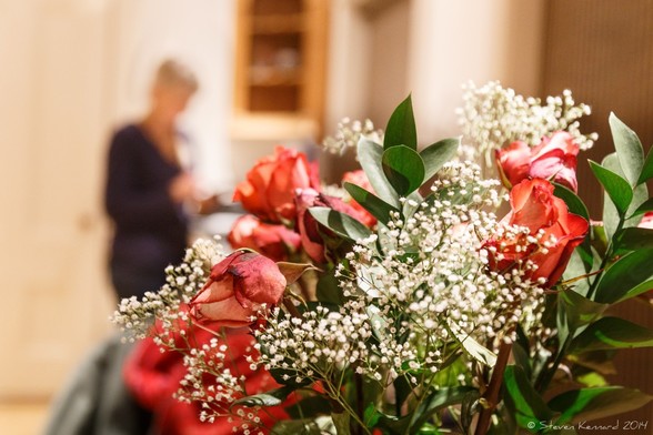 A bouquet of red roses, green leaves and white baby's breath (I think) in the foreground of a kitchen scene with my wife (out of focus) in the background.