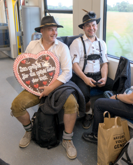 Photo of two men dressed for the Oktoberfest, riding a tram. One of them holds a large gingerbread heart with a German inscription: "the biggest heart for the biggest love of the world"
