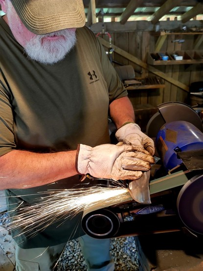 Man making and grinding a knife in a forge with sparks. 

Forge at Quiver full, Marrs Hill, North Carolina