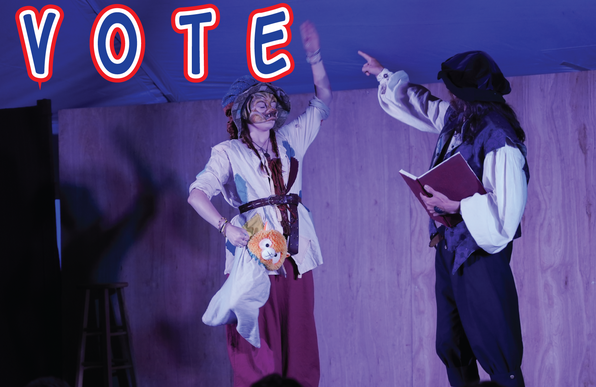 Two actors perform a commedia dell'arte play in a pavilion tent during a medieval camping event portraying the stock characters of Arlecchino taking instructions from il Dottore. The word "VOTE" in red, white, and blue is superimposed above them into the image.