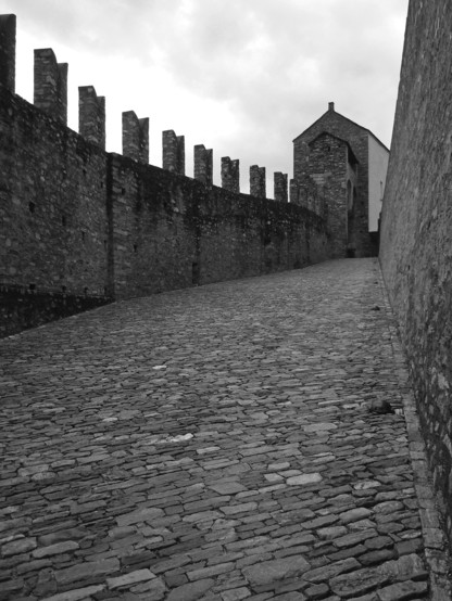 Black and white photo of a stretch of stone pavement that climbs up and to the right toward a three story structure and is bordered on the left by a tall, crenellated stone wall