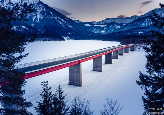 This stunning image captures an incredible bridge which arcs gracefully over a lake that is partially frozen with a generous coating of snow. It is set against a striking blue sky and breathtaking mountain range, which adds a majestic and awe-inspiring backdrop to the scene. The bridge itself is composed of a rectangular blue body with a red stripe on the side, and a railing running its length. On either side of the bridge, there are trees with white snow decorating their branches. On the left, an individual tree stands near the shore, while on the right, a group of trees appear to have been blanketed in snow. A hint of buildings can be seen in the background as well, with one having a tall red pole. This image is a perfect representation of the beauty of winter and the great outdoors.
