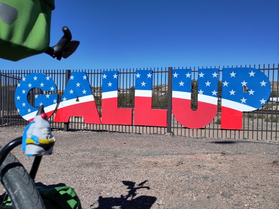 The Java Mascot "Duke" in front of a sign with the name of the city "Gallup". The letters are in the colors of the American flags and have stars on it.