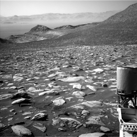 A black-and-white image shows a very rocky landscape of Mars from the viewpoint of the Curiosity rover. In the far background is a haze that covers up some of hills and sandy terrain. Closer to the rover, the sandy Martian surface is littered with smooth and bumpy rocks that slope down along a hill toward the far left. There are some wheel tracks closer to the foreground. On the right side of the image is part of the rover's body, a piece that is oval-shaped sits along the backside of the rover.