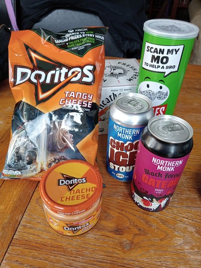 A pile of snacks, including a 4 pack of hazy IPA, 2 cans of stout, a tube of sour cream & chives flavoured Pringles, a big bag of tangy cheese Doritos and a jar of tangy cheese Dorito dip ðŸ˜‹