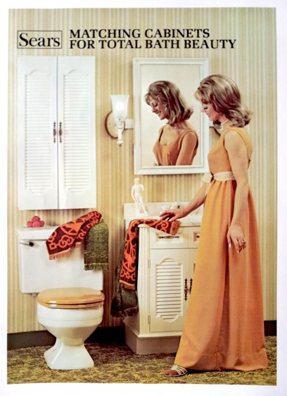 Female model with blonde styled hair wears orange formal gown. She touches a towel arranged artfully on a sink and vanity next to a toilet and a matching storage cabinet mounted above the toilet. A mirror reflects the model's head, showing her right profile. The scene is an advertising photo for Sears & Roebuck, circa 1978.