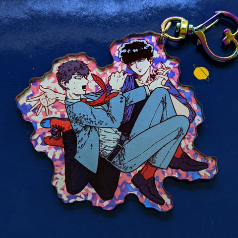 A photo of a charm made of see-through acrylic with a solid silver coating as background color that gives the color design printed on the charm a shining finish. The design is of two characters floating together, a man in an office suit and a man in casual wear. The man in casual wear is making the pair float with supernatural powers, creating a brilliant aura around them, while the man in the office suit holds on to him nervously.

The charm has a heart-shaped clasp.